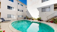 Located in the desirable Brentwood neighborhood of Los Angeles, this outdoor courtyard space centers around a kidney-shaped swimming pool that serves as its defining feature. The pool's clear turquoise water is complemented by a wide concrete pool deck, providing ample space for lounging and relaxation under the Southern California sun.

The courtyard is framed by a multi-story white stucco building on multiple sides, creating an intimate, enclosed atmosphere while still allowing for abundant natural light throughout the day. A metal staircase connects the pool level to upper-floor balconies, one of which features outdoor seating with a table and umbrella — ideal for al fresco dining with a view of the pool below.

Lush landscaping adds a welcoming, resort-like quality to the space. An assortment of potted plants, succulents, and tropical greenery lines the perimeter of the deck and pool area, introducing vibrant natural texture against the clean white exterior walls. Brick-bordered garden beds along the building's base further enhance the greenery.

The pool is equipped with metal handrail entry steps for easy access, and the surrounding deck provides space for lounge chairs. The courtyard's overall design balances practicality with comfort, offering residents a private outdoor retreat in the heart of West Los Angeles.