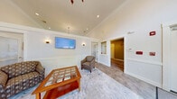 Located at 250 Skyline Dr in Dracut, MA, this two-floor community amenity space offers a generous and thoughtfully designed layout across both levels. The main floor hosts an elegant combined dining and living room distinguished by soaring ceilings, a dramatic arched transom window that floods the space with natural light, and French doors opening to an outdoor balcony with views of the surrounding landscape. Patterned carpeting, wainscoting, and warm chandelier lighting lend the room a refined, welcoming ambiance. Dining tables with upholstered chairs provide ample seating, while a comfortable living area with cushioned armchairs, a wood coffee table, and a wall-mounted flat-screen TV creates an inviting lounge setting. Hallways connecting the various spaces are tastefully finished with wainscoting, accent lighting, and decorative artwork. The spacious exercise room is comprehensively equipped with cardio machines including treadmills, ellipticals, and stationary bikes, as well as a variety of strength-training equipment, dumbbells, and a cable machine. Recessed lighting and neutral walls maintain a clean, functional atmosphere throughout the fitness area. Two bathrooms and dual staircases serve both floors, ensuring convenient access and circulation throughout this well-rounded facility.
