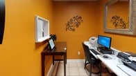 This single-room business center is designed to provide essential productivity amenities in a compact, functional layout. The space is immediately distinguished by its bold, warm orange walls, which lend an energetic ambiance to the room. Light-toned ceramic tile flooring runs throughout, contributing to a clean and professional appearance.

The workstation area serves as the focal point of the room, featuring a desktop computer with a monitor, keyboard, and mouse positioned on a white desk surface. Two printers — one laser and one multifunction — flank the computer, providing convenient printing and scanning capabilities. A decorative silver-framed mirror is mounted above the desk, adding a touch of elegance to the functional setup.

A secondary wooden side table offers additional surface space and displays a framed WiFi password sign alongside a decorative plant, ensuring guests can easily connect to the internet. A small interior window opening is situated above this table, adding architectural interest to the space.

The room is accessed through a classic six-panel white door with brass hardware. Wall-mounted dispensers and informational panels are positioned near the entrance, completing the space's practical, guest-oriented design.