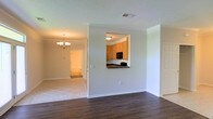 This single-story space presents a thoughtfully designed interior that balances open living with defined functional zones. The main living area features rich, dark hardwood flooring that anchors the space, while neutral light gray walls create a clean, versatile backdrop throughout. A rectangular pass-through window connects the living area to the kitchen, providing an open visual link between the two spaces while maintaining a sense of separation.

The kitchen, visible through the pass-through, is outfitted with warm honey-toned cabinetry, dark countertops, and black appliances, including a built-in microwave. A ceiling-mounted light fixture illuminates the kitchen efficiently.

Transitioning into the adjacent dining or secondary living area, the flooring shifts to light ceramic tile, adding textural contrast. A traditional-style chandelier with warm-toned bulbs serves as a decorative focal point, lending a touch of elegance to the space. A hallway beyond leads to additional rooms, suggesting a well-organized floor plan. White baseboards and door trim throughout provide crisp architectural definition, contributing to the overall clean and move-in-ready presentation of the interior.
