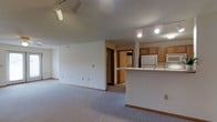 This single-floor space offers a clean, functional layout centered around an open-concept design that seamlessly connects the living area to the kitchen. The spacious carpeted living room is bathed in light and features track lighting mounted on the ceiling, providing versatile illumination throughout. Neutral white walls and wood-toned baseboards create a simple, understated aesthetic throughout the space.

The kitchen is positioned to one side and features warm oak cabinetry, a white refrigerator, a white microwave, and a peninsula with a light-colored countertop that doubles as a breakfast bar or prep area. The peninsula serves as a natural divider between the kitchen and living space while maintaining an open, airy feel. Track lighting continues into the kitchen, ensuring well-lit work surfaces.

A hallway visible from the main living area leads to additional rooms, hinting at further private spaces beyond the open floor plan. The textured ceiling adds subtle character to the interior. Overall, the space presents a practical and comfortable environment, well-suited for everyday living with its efficient layout, ample storage in the kitchen, and open flow between functional areas.