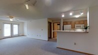 This single-floor space offers a clean, functional layout centered around an open-concept design that seamlessly connects the living area to the kitchen. The spacious carpeted living room is bathed in light and features track lighting mounted on the ceiling, providing versatile illumination throughout. Neutral white walls and wood-toned baseboards create a simple, understated aesthetic throughout the space.

The kitchen is positioned to one side and features warm oak cabinetry, a white refrigerator, a white microwave, and a peninsula with a light-colored countertop that doubles as a breakfast bar or prep area. The peninsula serves as a natural divider between the kitchen and living space while maintaining an open, airy feel. Track lighting continues into the kitchen, ensuring well-lit work surfaces.

A hallway visible from the main living area leads to additional rooms, hinting at further private spaces beyond the open floor plan. The textured ceiling adds subtle character to the interior. Overall, the space presents a practical and comfortable environment, well-suited for everyday living with its efficient layout, ample storage in the kitchen, and open flow between functional areas.