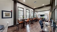 Located at 225 Sunlight Ln in Lake Mary, Florida, this sophisticated single-floor community or leasing lounge space delivers a polished, upscale atmosphere designed for both comfort and functionality. The interior is defined by rich hardwood-style flooring — including a striking herringbone-patterned section — complemented by bold dark wood trim, columns, and crown molding that contrast elegantly against crisp white walls.

The open layout is thoughtfully organized into distinct functional zones. A café-style dining area features tall bistro tables paired with leather-upholstered bar chairs, set along a generous row of large, dark-framed windows that flood the space with natural light and provide views of the lush outdoor greenery. A lounge area offers plush seating arrangements, including accent chairs and sofas, centered around a wall-mounted flat-screen television and a credenza with decorative art. Additional intimate seating nooks feature round tables with upholstered chairs, ideal for small gatherings or meetings.

Recessed lighting and pendant fixtures illuminate the space evenly, while interior windows and glass panel walls create visual connectivity between zones. Curated artwork, decorative vases, and greenery add warmth throughout. A wet bar or service counter with mosaic tile accents anchors one end of the space, enhancing its versatility. The overall ambiance is refined, welcoming, and professionally appointed.