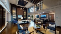 This single-story clubroom in Houston, TX blends industrial character with upscale residential design across several distinct functional zones. The centerpiece of the space is a dramatic double-height great room, where soaring ceilings with dark exposed beams draw the eye upward past an expansive grid of steel-framed clerestory windows that flood the interior with natural light. A full-height exposed brick accent wall anchors one end of the room, featuring a built-in fireplace, a wall-mounted television, and flanking illuminated shelving with dark cabinetry—creating a cozy yet striking focal point. Multiple seating arrangements populate the main lounge, including tufted sofas, leather chairs, and blue upholstered accent pieces arranged around marble-topped coffee tables, encouraging both lively gatherings and quiet relaxation. Dark hardwood flooring runs throughout, grounding the space with warmth. A dedicated catering kitchen offers dark cabinetry, grey stone countertops, stainless steel appliances, and a wall-mounted display screen. A separate private dining area accommodates a large party around a long wood table, set against another exposed brick wall adorned with curated artwork and framed by steel-framed windows overlooking the outdoors. An additional flex area with light tile flooring and a mounted television provides versatile space for various activities.