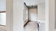 This one-bedroom, two-bathroom unit blends industrial character with modern finishes across a single, efficiently organized floor. The living room serves as the heart of the space, showcasing a striking combination of whitewashed and natural exposed brick walls, track lighting, and hardwood-style flooring that flows throughout. An arched brick detail and large windows bring in natural light, enhancing the loft-inspired aesthetic.

The kitchen is well-appointed with white cabinetry, gray countertops, and a full suite of stainless steel appliances including a gas range, microwave, refrigerator, and dishwasher. Open industrial-style shelving adds both function and visual interest, while a kitchen island with an undermount sink provides additional prep space. A doorway from the kitchen leads directly into a dedicated walk-in closet with built-in shelving.

The bedroom offers a clean, versatile layout with exposed structural elements overhead and ample wall space. Two bathrooms serve the unit — one features a wood-toned vanity, natural light from a window, and painted brick walls, while the second includes a wood-panel tiled shower surround and a matching vanity. In-unit washer and dryer hookups complete this thoughtfully designed urban residence.