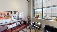 Located at 30–98 Market St in Camden, NJ, this urban loft delivers a striking blend of industrial character and contemporary comfort. The space is defined by its dramatically tall ceilings, exposed ductwork, and bold track lighting that runs along ceiling-mounted rails, all hallmarks of its converted warehouse origins.

The living area is bright and expansive, anchored by large, multi-pane factory-style windows with teal-framed casings that flood the room with natural light and offer views of the surrounding cityscape and tree canopy. The open layout flows naturally between the living and sleeping areas.

The kitchen features white cabinetry, granite-style countertops, a full appliance suite, and tile flooring, offering a clean, functional workspace. The bedroom areas continue the loft aesthetic with soaring ceilings, track lighting, and generous windows. A walk-in closet provides ample storage. Two full bathrooms are included — one with a pedestal sink and tub/shower combo, and another with a full tub/shower — both finished in neutral tones with tile flooring.

Throughout the space, the interplay of raw architectural elements and clean white walls creates an airy, gallery-like ambiance well-suited for urban living in the heart of Camden.