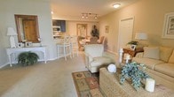 This single-floor residence offers an open-concept layout that seamlessly connects the living, dining, and kitchen areas into one cohesive, functional space. The living area is furnished with a comfortable upholstered armchair and a woven storage ottoman, grounded by a nature-themed area rug featuring coastal motifs. Decorative accents, including candle stands, framed photos, and lush greenery, contribute to a warm, relaxed atmosphere.

The dining area sits just beyond the living space, featuring a round table with wicker-style chairs and is enhanced by a tropical botanical artwork on the wall, reinforcing the coastal aesthetic. A tall, lush indoor plant adds a natural, organic element to the corner.

The kitchen is partially open to the main living area, separated by a raised breakfast bar with white bar stools. The kitchen features dark cabinetry, stainless steel appliances including a microwave and refrigerator, and light countertops. Track lighting above the bar area provides focused illumination.

Neutral beige carpeting and walls flow throughout, creating a cohesive and calming environment. The layout is efficient and well-lit, offering a comfortable, easy-to-navigate single-level living experience.