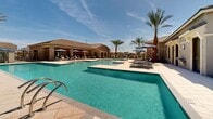 Located at 6900 S 59th Ave in Phoenix, Arizona, this single-story community amenity center offers a resort-inspired outdoor experience designed for relaxation and recreation. The centerpiece of the space is a generously sized swimming pool with crystal-clear turquoise water, complemented by a separate shallow-entry pool and spa, all finished with clean tile surrounds and wide, light-toned paver decking. Ample lounge chairs and shaded umbrellas in coordinating navy blue are arranged throughout the sun deck, providing plenty of space for sunbathing or unwinding poolside. Lush turf areas with additional lounge seating add a touch of greenery to the desert landscape, while mature palm trees frame the area and enhance the warm Arizona atmosphere. The surrounding architecture features a stucco exterior with pitched tile rooflines consistent with the Southwest aesthetic of the broader community. A dedicated fitness studio, operated under the Yardly brand, is accessible directly from the pool deck, offering residents convenient access to workout facilities. The overall ambiance is bright, open, and inviting, taking full advantage of the abundant Phoenix sunshine and clear blue skies that define the region's climate year-round.