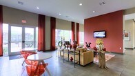 This well-appointed single-floor space makes a striking first impression with its soaring ceilings and vibrant design palette. The open layout is anchored by a bold terracotta-red accent wall, which pairs seamlessly with deep crimson floor-to-ceiling curtains framing the large windows that flood the interior with abundant natural light and views of lush greenery outside.

The main gathering area features a comfortable tan upholstered sofa set arranged around a decorative open-frame shelving unit adorned with curated ceramic vases, candles, and sculptural accents in warm earthy tones. A wall-mounted flat-screen television is positioned prominently on the red accent wall, making the space ideal for both relaxing and socializing. A rustic natural wood side table adds an organic contrast to the otherwise contemporary aesthetic.

Near the windows, a casual dining or conversation nook is set with a round white table and modern transparent red chairs, adding a pop of color and playful energy to the space. The large-format tile flooring throughout lends a clean, polished finish that ties the entire design together.

Recessed ceiling lighting complements the natural light, ensuring the space remains bright and inviting at any hour. Overall, the interior exudes a warm, modern ambiance with a cohesive blend of bold color, texture, and thoughtful décor.