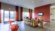 This well-appointed single-floor space makes a striking first impression with its soaring ceilings and vibrant design palette. The open layout is anchored by a bold terracotta-red accent wall, which pairs seamlessly with deep crimson floor-to-ceiling curtains framing the large windows that flood the interior with abundant natural light and views of lush greenery outside.

The main gathering area features a comfortable tan upholstered sofa set arranged around a decorative open-frame shelving unit adorned with curated ceramic vases, candles, and sculptural accents in warm earthy tones. A wall-mounted flat-screen television is positioned prominently on the red accent wall, making the space ideal for both relaxing and socializing. A rustic natural wood side table adds an organic contrast to the otherwise contemporary aesthetic.

Near the windows, a casual dining or conversation nook is set with a round white table and modern transparent red chairs, adding a pop of color and playful energy to the space. The large-format tile flooring throughout lends a clean, polished finish that ties the entire design together.

Recessed ceiling lighting complements the natural light, ensuring the space remains bright and inviting at any hour. Overall, the interior exudes a warm, modern ambiance with a cohesive blend of bold color, texture, and thoughtful décor.