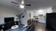 This single-story space showcases an open-concept design that integrates the living, dining, and kitchen areas into one cohesive, functional layout. The living area is anchored by a plush gray sofa, a round white coffee table, and a patterned blue area rug, with a ceiling fan with integrated lighting overhead providing both airflow and ambient illumination. A large tropical plant adds a natural, organic element to the corner of the room, complementing the clean, neutral wall tones throughout.

The dining area transitions naturally from the living space, featuring a compact table with leather-accented chairs, ideal for intimate meals. This flows directly into the kitchen, which is outfitted with crisp white cabinetry, a tile backsplash, and stainless steel appliances, including a dishwasher and sink positioned beneath upper cabinets for practical storage. The countertops are clean and uncluttered, reinforcing an efficient use of space.

Hardwood-style flooring runs throughout the main living areas, adding warmth and cohesion to the interior. The overall ambiance is modern and well-maintained, with a bright, airy feel supported by thoughtful lighting and a neutral color palette that enhances the sense of openness.