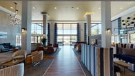 This contemporary amenity center is defined by its dramatic double-height lobby, where soaring ceilings, floor-to-ceiling windows, and a golden accent ceiling create an immediately impressive entrance. Expansive glazing floods the interior with natural light and offers unobstructed views of the surrounding outdoor courtyard and greenery. The main lounge features herringbone-patterned hardwood flooring, a sleek linear fireplace set against a textured stone accent wall, and a striking geometric chandelier overhead — all anchored by curated seating arrangements with leather and upholstered chairs. Additional lounge areas are thoughtfully arranged throughout, offering intimate gathering spots with stylish mixed-material furniture and large-format TVs. A dedicated bar and kitchen area includes professional-grade stainless steel appliances, a white quartz countertop island, and bar seating with marble-tiled flooring, ideal for entertaining. A separate dining lounge with a large communal table surrounded by teal upholstered chairs provides a versatile space for group events. The outdoor covered patio features bold blue sectional seating and opens directly to a shared courtyard, framed by exposed brick and floor-to-ceiling glass sliders that blur the boundary between indoors and out.