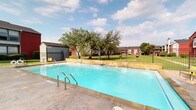 This residential community offers a blend of comfort and amenities in a suburban setting. The property is characterized by its distinctive red and white exterior buildings, creating a visually appealing contrast against the lush green landscape. A standout feature is the large, rectangular swimming pool, complete with depth markers and sturdy metal railings for safety. The pool area is surrounded by a concrete deck, perfect for lounging or sunbathing.

The grounds are meticulously maintained, with well-manicured lawns and strategically placed trees providing natural shade. A black metal fence encircles the property, offering both security and aesthetic appeal. The complex includes multiple buildings, suggesting a variety of apartment sizes and layouts to accommodate different needs.

A covered outdoor common area is visible, featuring a wooden deck and protective railing. This space is nestled under the canopy of a large, mature tree, creating a serene spot for residents to gather or relax. The overall design of the complex emphasizes open spaces and greenery, fostering a sense of community while maintaining individual privacy. The architecture combines modern elements with traditional touches, resulting in a welcoming and attractive living environment.