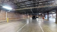 This single-level indoor parking facility offers a generous, open layout designed to accommodate a substantial number of vehicles with ease. The space is defined by its striking exposed brick walls, which lend an industrial character and hint at the building's historic construction. Overhead, a robust steel truss and metal deck ceiling system spans the full width of the facility, providing both structural integrity and considerable vertical clearance. The floor is finished in durable concrete, with painted lines delineating individual parking stalls in an organized grid pattern. Lighting fixtures are mounted throughout the ceiling structure, ensuring adequate illumination across the entire parking area. Support columns are positioned at regular intervals, and a yellow safety bollard is visible near one of the walls, reflecting attention to safety and traffic flow. The overall ambiance is utilitarian yet well-maintained, with the combination of masonry walls and steel framework creating a sturdy, long-lasting environment. This facility is well-suited for both everyday parking needs and larger-volume vehicle storage, offering a secure and covered alternative to surface parking.