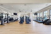 Located at Yardly at Sundance in Buckeye, Arizona, this well-appointed fitness center offers a generously sized, single-floor layout designed to support a full range of workout routines. The space features light hardwood-style flooring throughout, crisp white walls, and a tray ceiling accented by multiple ceiling fans that keep the environment comfortable. Recessed lighting complements the abundant natural light streaming through large, black-framed windows, many of which frame views of the outdoor pool area and surrounding palm trees, creating an energizing atmosphere.

The main workout area is comprehensively equipped with a row of treadmills, multi-station cable and strength machines, a dumbbell rack with adjustable bench, TRX suspension trainers, and a functional training rig. A dedicated cardio section houses multiple stationary cycling bikes and elliptical and rowing machines, each featuring individual screens for interactive workouts. Wall-mounted flat-screen televisions are positioned throughout for added entertainment. The Yardly branding is prominently displayed on mirrored wall panels, reinforcing the community identity. Glass double doors provide direct access to the outdoor amenity areas, seamlessly connecting indoor fitness with the outdoor lifestyle that defines this Buckeye community.
