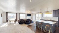 This well-appointed single-floor space offers a seamlessly integrated kitchen and living area designed with contemporary style and functionality in mind. The open-concept layout allows for effortless flow between the two zones, unified by large-format dark tile flooring in the kitchen area and plush carpeting throughout the living space.

The kitchen is a standout feature, showcasing dark espresso cabinetry, light quartz countertops, a mosaic tile backsplash, and stainless steel appliances including a French door refrigerator. A large central island with seating for multiple guests serves as both a prep space and a casual gathering point, illuminated overhead by a pair of sleek dome pendant lights.

The living area is bright and inviting, benefiting from abundant natural light streaming through multiple large windows and glass French doors that open to an outdoor balcony with sweeping urban and mountain views. A clean, white-surround fireplace anchors the seating area, complemented by a tray ceiling with a statement Sputnik chandelier and recessed lighting. A dining area with a table and chairs occupies a corner of the room.

A separate game room rounds out the space, featuring a striking geometric shuffleboard table, carpeted flooring, and a casual bulletin board wall, making it ideal for recreation and socializing.