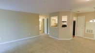 This single-floor residence is designed with an open and functional layout that maximizes the sense of space throughout. The main living area is finished with plush wall-to-wall carpeting in a warm neutral tone, complemented by smooth white ceilings and clean white baseboards that create a bright, cohesive aesthetic. Walls are painted in a soft, muted beige-green palette that lends a calm and welcoming ambiance to the interior.

A defining architectural feature is the pass-through opening connecting the living area to the kitchen, which enhances the open feel and allows for easy interaction between spaces. The kitchen is visible through this cutout and features white cabinetry, a sink, and black appliances, offering a practical and tidy cooking environment. A second, smaller pass-through or serving window is also visible nearby, adding further connectivity.

A white paneled door leads from the main living space toward additional rooms, including what appears to be a hallway with access to further areas of the unit. Natural light filters in from adjacent spaces, brightening the interior without the need for heavy artificial lighting. The overall design is clean, understated, and move-in ready, with a straightforward layout well-suited for comfortable everyday living.