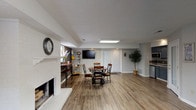 This single-floor leasing center offers a functional and inviting environment designed to welcome prospective residents. The space is finished with warm wood-look flooring throughout, creating a cohesive and polished aesthetic across all areas.

The main gathering area serves as the heart of the leasing center, featuring a white brick fireplace with a classic mantel as a prominent focal point. A comfortable sofa positioned nearby creates a relaxed lounge setting, while a wall-mounted flat-screen television adds a modern touch. A large overhead skylight panel floods the central space with bright, even light.

Adjacent to the lounge, a round dining table with wrought-iron chairs provides a functional meeting area, ideal for consultations or community events. Double French doors with frosted glass panels and horizontal blinds offer a private entry point and allow natural light to filter in.

A compact kitchenette is neatly tucked into an alcove, outfitted with slate-blue cabinetry, stone-look countertops, a stainless steel sink, microwave, and refrigerator—providing convenient amenities for staff and visitors alike.

A private office area is set apart from the main floor, furnished with a full desk and shelving to support leasing operations. The overall color palette of soft grays, warm whites, and charcoal accent walls gives the space a professional yet approachable ambiance.