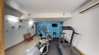 This fitness room offers a practical and functional workout space equipped with everything needed for a solid cardio session. The room features multiple elliptical machines and a treadmill, providing a variety of options for cardiovascular exercise. A wall-mounted flat-screen television is positioned against a bold blue accent wall, allowing guests or residents to stay entertained during their workouts. A classic wooden bench near the entrance provides a convenient spot for stretching or resting between sets. The space is finished with a drop ceiling fitted with recessed lighting, ensuring the room is well-lit throughout. Durable, dark rubberized flooring covers the workout area, offering the grip and cushioning appropriate for exercise equipment, while light tile flooring defines the entry area. A wall clock keeps exercisers on schedule, and a phone mounted near the entrance adds a practical touch. The overall layout is efficient and straightforward, making good use of the available space to accommodate the core fitness needs of its users.