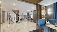 Located at 100 Waddell Plaza Lane in Cary, NC, this expansive two-story amenity space offers a compelling blend of modern architecture and upscale interior design. The ground floor makes an immediate impression with a dramatic double-height lobby showcasing soaring ceilings, large-scale textured stone wall panels, and an eye-catching chandelier installation. Floor-to-ceiling windows flood the space with natural light and provide views of the surrounding exterior. Polished marble-look tile flooring with gold veining flows seamlessly through the entrance and main living areas, while a patterned commercial carpet defines the lounge zones furnished with contemporary seating arrangements. A sweeping curved staircase with glass railings connects the two floors, complemented by an elevator for accessibility. The upper level features a hallway with decorative wall art and additional seating. The second floor also opens to a mezzanine overlook. A dedicated game and entertainment lounge includes a full-size pool table, a built-in wet bar with dark subway tile backsplash, a large-screen TV, and a tufted blue banquette seating area beneath elegant pendant lighting. Gold-toned accents, slatted wall panels, and curated artwork throughout unify the space with a refined, contemporary aesthetic.