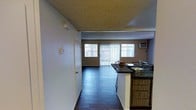This two-floor residence offers a thoughtfully designed open-concept layout that seamlessly connects the kitchen and living areas. The kitchen features a raised breakfast bar with dark granite countertops and warm wood cabinetry, providing both functional workspace and a casual dining option. Dark hardwood flooring runs throughout the main level, adding a rich, cohesive feel to the interior. The textured popcorn ceiling adds a classic touch overhead, while the smooth walls are painted in neutral tones that complement the overall aesthetic.

The living area is bright and welcoming, benefiting from a large window and sliding glass door dressed with vertical blinds that allow for adjustable natural light. A wall-mounted air conditioning unit ensures year-round comfort. The open sightlines from the kitchen through to the living space create an expansive, airy atmosphere well-suited for both everyday living and entertaining.

A hallway door off the main area provides access to additional rooms, contributing to a practical and well-organized floor plan. Spanning two floors, this residence offers ample room to accommodate a variety of living arrangements, with the main level establishing a strong foundation of functionality and comfort.