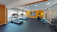 This single-story fitness center in Jacksonville, Florida, is designed to provide a functional and energizing workout environment. The space is divided into two connected areas, unified by a bold orange accent wall that adds a vibrant, motivating atmosphere throughout. A large pass-through window between the two sections maintains visual openness and allows natural light to flow freely across both areas.

The main cardio zone features multiple treadmills positioned along a wall of large windows that overlook an outdoor pool area, offering pleasant views and ample natural light during workouts. An elliptical trainer and stationary spin bike complement the cardio equipment selection. Durable dark rubber flooring covers the entire space, providing both safety and sound absorption.

The adjacent free weights area houses a well-stocked dumbbell rack with a wall-mounted mirror, allowing users to monitor their form. A water cooler station, seating bench, and waste bin are conveniently placed near the entry, adding practical amenities. The space is illuminated by a combination of recessed ceiling lights and fluorescent fixtures beneath a textured ceiling. French doors lead directly outside, enhancing ventilation and reinforcing the connection to the surrounding community.