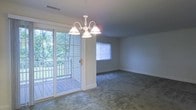 This single-floor residence offers a comfortable and well-lit living environment defined by classic design details and functional layout. The main living area is finished with wall-to-wall carpeting in a neutral tone, lending a warm and cozy feel to the space. A white chandelier with elegant curved arms serves as a central decorative focal point, casting a soft, inviting glow throughout the room.

Natural light flows generously into the space through multiple windows fitted with white blinds, as well as through a set of sliding glass doors framed by plantation shutters. These doors open directly onto a wood deck that overlooks lush, mature greenery, creating a seamless connection between the interior and the outdoors. The deck provides a pleasant extension of the living space, ideal for relaxing or entertaining while enjoying the natural surroundings.

The room's neutral gray-toned walls and white trim offer a clean, understated backdrop that complements a variety of furnishing styles. Ceiling-mounted ventilation is integrated discreetly into the layout. Overall, the space strikes a balance between classic residential charm and everyday practicality, making it a welcoming and versatile environment throughout the single level.