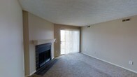 This single-floor residence offers a functional and comfortable layout designed for everyday living. The living area serves as the heart of the space, anchored by a classic fireplace with a white painted mantel and dark tile surround, providing a warm and inviting focal point. Plush wall-to-wall carpeting runs throughout the living area, adding comfort underfoot and a neutral aesthetic that complements the soft, beige-toned walls. Natural light flows generously into the room through a full-height sliding glass door dressed with vertical blinds, which also provides access to an outdoor area, seamlessly connecting interior and exterior living. The textured ceiling adds a subtle architectural detail, while the clean white baseboards frame the space neatly. Ceiling vents are integrated discreetly, ensuring climate comfort throughout. The overall ambiance of the space is calm, understated, and move-in ready, with a straightforward design that offers a solid canvas for personalization. The single-floor layout ensures ease of navigation, making the space practical and accessible for a variety of lifestyles.