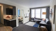 This single-floor unit offers a thoughtfully arranged layout that makes efficient use of its available space. The living area serves as the central hub, featuring dark carpeting that contrasts with crisp white walls and bright white crown molding that adds a classic architectural touch. A large set of windows along one wall, framed by a dark charcoal accent wall, allows generous natural light to fill the room, creating an open and airy atmosphere. The living space accommodates a seating arrangement anchored by layered area rugs, while a wall-mounted flat-screen television provides a focal point opposite the sofa.

A doorway from the living area leads directly into the bedroom, which continues the dark carpeting and features a blue accent wall, adding a pop of color. The bedroom appears to comfortably fit a full-sized bed with additional furnishings. Adjacent to the main living space, a kitchenette is visible, equipped with dark cabinetry, a sink, and counter space, providing essential cooking and prep functionality. The overall ambiance of the unit is modern and functional, balancing contrast through dark and light tones throughout.