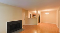 This single-floor space is designed with an open-concept layout that seamlessly connects the living area to the kitchen. The living room is a welcoming space anchored by a classic fireplace with a dark tile surround and hearth, adding both warmth and a focal point to the room. Light hardwood flooring extends throughout, contributing to a bright and cohesive aesthetic.

The kitchen is accessible via a raised pass-through counter, which creates a natural division between the cooking and living areas while maintaining an open feel. Warm wood cabinetry lines the kitchen walls, complemented by stainless steel appliances, including a microwave and refrigerator, as well as a range. White upper cabinetry with glass-panel doors adds a subtle contrast and additional storage.

The walls throughout are finished in a soft, neutral warm tone that enhances the overall brightness of the space. White trim and baseboards provide clean definition along the floors and doorways. A chandelier is visible in an adjoining area, suggesting additional living or dining space. The single-floor design ensures ease of movement and accessibility throughout.