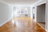 Located at 83-33 118th St in the charming Kew Gardens neighborhood of Queens, New York, this single-floor residence offers a thoughtfully designed layout with a clean, neutral aesthetic throughout. Warm-toned parquet wood flooring is a defining feature found across the living, dining, and bedroom areas, adding timeless character to every room.

The spacious living area is bathed in natural light from a large multi-pane window and features crown molding, soft gray walls, and a wall-mounted air conditioning unit paired with a classic radiator for year-round comfort. The open sightlines create an airy, welcoming atmosphere.

The bedroom continues the parquet flooring and includes a sliding closet with built-in shelving, providing ample storage. A window brings in natural light, completing the comfortable and functional space.

The kitchen has been updated with gray shaker-style cabinetry, dark countertops, white subway tile backsplash, hardwood-style flooring, and stainless steel appliances including a gas range and built-in microwave.

The bathroom is finished with white tile, a modern vanity with a mirrored medicine cabinet, and a tub-shower combination with a sliding glass enclosure. Every room reflects a practical and well-maintained living environment ideal for urban living.