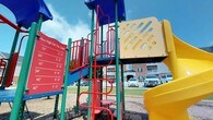 Spring Creek's outdoor playground is a lively and well-equipped recreational space designed to engage children of all ages. The centerpiece of the area is a multi-component play structure featuring bold primary colors — vivid greens, reds, yellows, and blues — that make the space visually dynamic and inviting. The structure includes climbing ladders, a spiral climber, enclosed tube slides, and an interactive "How High Can You Jump?" measurement panel, encouraging both physical activity and friendly competition among young visitors. The playground surface is finished with a wood chip base, providing a safe and cushioned ground covering beneath the equipment. Surrounding the play area is a well-maintained open lawn that offers additional space for outdoor recreation. Mature trees line the perimeter, providing natural shade on sunny days. In the background, the residential buildings of Spring Creek are visible, featuring classic brick and neutral-toned facades with covered entryways and balconies, reflecting a cohesive and carefully planned community aesthetic. The open sky and natural light contribute to an upbeat, cheerful atmosphere, making this playground a standout amenity within the Spring Creek community.