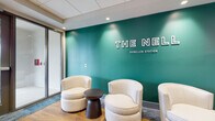 The Nel at Dunellen Station presents a polished and professionally designed lobby that serves as the welcoming hub of the building. The space immediately draws attention with a striking teal accent wall bearing the property's name and "Dunellen Station" branding in bold white lettering, creating a strong visual identity from the moment of arrival.

The lobby's flooring transitions seamlessly between warm wood-tone planks and textured blue-grey carpet, adding visual depth and defining distinct zones within the space. Comfortable cream-upholstered swivel chairs are arranged around a dark round side table, offering residents and visitors an inviting lounge area ideal for casual conversation or a brief rest.

Recessed lighting throughout the drop ceiling provides consistent, well-distributed illumination, enhancing the clean and contemporary aesthetic. A stainless steel elevator with modern call panels is centrally accessible, while a large wall-mounted display screen nearby adds a functional, modern touch to the corridor area.

Adjacent to the main entrance — which features glass double doors opening to the exterior — is a dedicated leasing office enclosed in glass, clearly signed and designed to be approachable and transparent. The overall ambiance of the lobby is sleek, welcoming, and brand-forward, reflecting a thoughtfully curated residential community experience at The Nel at Dunellen Station.