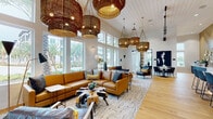 Located at 12231 Pioneers Way in Orlando, FL, this single-floor clubhouse is a thoughtfully designed community gathering space that blends contemporary style with welcoming comfort. The main lounge area commands attention with soaring ceilings, floor-to-ceiling windows, and an abundance of natural light that highlights the warm honey-toned hardwood floors. Oversized woven rattan pendant lights hang dramatically overhead, complementing plush caramel leather sectionals, layered area rugs, and curated accent pieces that give the space a refined yet inviting character.

A dedicated game room features a full-size pool table centered beneath a cluster of gold pendant lights and an ornate chandelier, with built-in banquette seating along the walls and ample windows offering views of the surrounding palm-lined landscape.

The kitchen and bar area is anchored by a long white countertop island with under-counter lighting, lined with steel-framed upholstered bar stools. Dark navy cabinetry, open floating shelves with decorative objects, a mounted television, and globe pendant lights contribute to the space's modern-industrial aesthetic.

A cozy dining nook features a round marble-top table, sleek upholstered chairs, and a decorative wall installation of wooden framed panels. The entry area is accented with bold black-and-white artwork and sculptural lighting, setting a sophisticated tone from the moment of arrival.