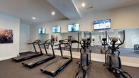 Located at 5793 Hoover Falls Dr in Westerville, Ohio, this fitness center offers a well-equipped, single-floor exercise facility designed to support an active lifestyle. The cardio area is lined with a row of modern treadmills and elliptical machines, each positioned in front of large, wall-mounted flat-screen televisions, allowing members to stay entertained during their workouts. Full-length mirrors span the length of the main workout wall, visually expanding the space and providing users with proper form feedback during exercise. The room features a neutral color palette with light gray walls and white trim, contributing to a clean and motivating atmosphere. Recessed lighting throughout the ceiling ensures the space is consistently bright and well-illuminated. The low-pile carpet flooring provides a practical and comfortable surface suited for fitness use. The overall design is functional yet polished, reflecting a commitment to providing residents with a quality fitness experience. The space is thoughtfully arranged to maximize usability while maintaining an open, uncluttered feel.