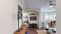 This single-story residence offers a thoughtfully designed layout across two bedrooms and three bathrooms. The open-concept living, dining, and kitchen area forms the heart of the home, anchored by rich hardwood-style flooring and crown molding details. The living room features a distinctive arched alcove housing a wall-mounted television, complemented by a long wooden console and curated artwork. Natural light fills the dining area through multiple windows dressed with plantation shutters and sheer curtains, framing outdoor greenery views around a rounded wood dining table.

The galley-style kitchen is equipped with stainless steel appliances, white upper cabinetry, gray lower cabinets, quartz countertops, and a tile backsplash. A dedicated laundry room with wire shelving provides added convenience. A central hallway connects both bedrooms, each finished with plush carpet and windows with outdoor views. The primary bedroom offers a ceiling fan and ample natural light. Both bedrooms reflect a calm, neutral palette. Three full bathrooms serve the layout efficiently. Throughout the residence, a cohesive blend of warm wood tones, neutral walls, and contemporary fixtures creates a refined, comfortable ambiance.