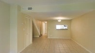 This two-story home offers a clean, functional layout designed for comfortable everyday living. The main level features a generously sized living area finished with light-toned ceramic tile flooring that runs throughout, lending a bright and cohesive feel to the space. Warm, neutral-painted walls complement the overall airy atmosphere, while ceiling-mounted light fixtures provide steady, even illumination.

A white-railed staircase rises from the main level, connecting to the upper floor and serving as a natural focal point within the open layout. Adjacent to the staircase, a louvered bifold closet door offers practical storage space. A notable architectural detail on the main level is a rectangular interior pass-through window, which connects the living area to what appears to be the kitchen, enhancing the flow of light and communication between spaces.

The hallway leading toward the rear of the home provides access to additional rooms, with a central ceiling-mounted light guiding the way. Overhead HVAC vents are integrated into the ceiling, ensuring climate comfort throughout. The overall design is simple and understated, with clean lines and a neutral palette that creates a versatile and welcoming environment suitable for a variety of interior arrangements.