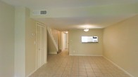 This two-story home offers a clean, functional layout designed for comfortable everyday living. The main level features a generously sized living area finished with light-toned ceramic tile flooring that runs throughout, lending a bright and cohesive feel to the space. Warm, neutral-painted walls complement the overall airy atmosphere, while ceiling-mounted light fixtures provide steady, even illumination.

A white-railed staircase rises from the main level, connecting to the upper floor and serving as a natural focal point within the open layout. Adjacent to the staircase, a louvered bifold closet door offers practical storage space. A notable architectural detail on the main level is a rectangular interior pass-through window, which connects the living area to what appears to be the kitchen, enhancing the flow of light and communication between spaces.

The hallway leading toward the rear of the home provides access to additional rooms, with a central ceiling-mounted light guiding the way. Overhead HVAC vents are integrated into the ceiling, ensuring climate comfort throughout. The overall design is simple and understated, with clean lines and a neutral palette that creates a versatile and welcoming environment suitable for a variety of interior arrangements.