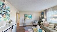 Located at 900 Plaza Rd in Atlantic Beach, Florida, this single-floor residence embraces a vibrant coastal aesthetic throughout. The living room serves as the heart of the home, featuring wood-look flooring, a textured ceiling, and large windows that invite natural light. Beach-inspired décor — including ocean-themed artwork and layered rugs — creates a relaxed, welcoming ambiance. A dedicated entryway with a coat rack provides a functional transition into the living space.

The kitchen is well-appointed with grey shaker-style cabinetry, granite countertops, stainless steel appliances including a range, microwave, and dishwasher, and wood-look tile flooring. A stacked washer and dryer unit is conveniently tucked into an adjacent closet.

The dining area offers seating for six beneath sheer curtain-draped windows, accented by nautical wall art that reinforces the coastal theme throughout.

The primary bedroom features plush carpeting, a distinctive blue reclaimed wood headboard, and ample dresser and storage space. The second bedroom includes dual closets and a bright window with sheer curtains. The bathroom is finished with granite countertops, white cabinetry, and tiled flooring with a shower-tub combination.
