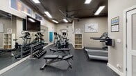 Located at 1646 River St in Des Plaines, Illinois, this fitness center offers a well-equipped, single-floor workout facility designed to support a full range of exercise routines. The space features durable rubber flooring throughout, providing a safe and functional surface for all types of training. Overhead fluorescent lighting and recessed fixtures ensure the room is brightly and evenly illuminated, while a ceiling fan promotes air circulation during workouts. Exposed ductwork along the ceiling gives the space an industrial character, complementing the neutral-toned walls accented with motivational fitness-themed typography and framed artwork. The cardio zone is outfitted with treadmills and elliptical machines, offering options for both walking and low-impact training. A dedicated free weights area includes a full dumbbell rack displayed alongside a large wall mirror, supporting strength training and form monitoring. An adjustable weight bench provides versatility for a variety of exercises. A multi-function cable machine, accompanied by stability and medicine balls, rounds out the strength training offerings. A small open shelving unit keeps essentials organized and accessible. The facility connects directly to a lobby area with a mail center, and signage indicates proper safety and usage guidelines throughout the space.