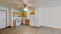 This single-floor space offers a practical and well-organized layout designed for comfortable everyday living. The kitchen is a standout feature, showcasing warm honey-toned wood cabinetry that lines the walls in an L-shaped configuration, providing generous storage throughout. The kitchen is fully equipped with a gas range, dishwasher, and a full-size refrigerator, all finished in white for a clean, cohesive look. A built-in sink with a window-side position adds functionality, while the under-cabinet range hood keeps the space practical. The kitchen floor features neutral tile that is both durable and easy to maintain. A ceiling fan with an integrated light fixture is centrally mounted, ensuring year-round air circulation and ambient lighting throughout the open area. The kitchen transitions naturally into the adjacent living space, which is finished with wall-to-wall carpeting, adding warmth and comfort underfoot. Neutral wall tones throughout create a bright, airy atmosphere that feels open and welcoming. The overall design emphasizes functionality and simplicity, making this a well-suited space for those seeking a straightforward, move-in-ready layout with all essential features in place.