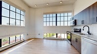 This thoughtfully designed single-floor unit blends industrial character with contemporary finishes throughout its five rooms. Soaring ceilings with exposed beams and ductwork lend an airy loft-like quality, while large factory-style multi-pane windows flood every room with abundant natural light and frame impressive city skyline views. Light-toned hardwood flooring runs continuously through the living areas and bedrooms, creating a warm, cohesive aesthetic.

The open-concept kitchen and living area serves as the heart of the unit, anchored by a full suite of stainless steel appliances, including a refrigerator, range, microwave, and dishwasher. Sleek wood-toned cabinetry with matte black hardware and a subway tile backsplash complete the modern kitchen aesthetic.

The bedrooms are bright and well-proportioned, each featuring high ceilings, large windows with city views, and ample double-door closet storage with matching black hardware. A hallway connects the rooms with clean white walls and recessed lighting.

The bathroom is finished to a high standard, offering a wood-grain vanity with a white quartz countertop, a matte black faucet, and a frameless glass walk-in shower with white subway tile surround and a linear drain. The overall ambiance throughout is clean, modern, and bright, with a nod to the building's industrial heritage.