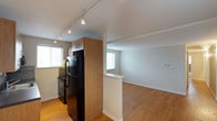 This single-floor space showcases an open-concept layout that connects the kitchen and living area seamlessly. The kitchen is equipped with black appliances, including a full-size refrigerator and a range, complemented by warm wood-toned cabinetry that adds a natural element to the space. A track lighting system runs along the white ceiling, providing adjustable and functional illumination throughout the kitchen area. A partial wall divider subtly defines the transition between the kitchen and the adjoining living space, maintaining an open and airy feel without fully separating the two areas. The living area features light hardwood-style flooring that extends throughout, contributing to a cohesive and inviting atmosphere. Multiple windows fitted with horizontal blinds allow natural light to filter in, brightening the interior and enhancing the sense of openness. The neutral-toned walls throughout provide a clean, versatile backdrop suitable for a variety of interior styles. White baseboards and trim offer a crisp finishing detail that ties the overall aesthetic together. The layout is practical and well-organized, making efficient use of the available space.