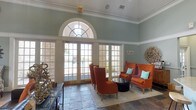 This single-story lounge space makes a strong architectural impression with its notably high ceilings adorned with elegant crown molding. The focal point of the room is a grand arched transom window positioned above a set of white-framed French doors, flooding the interior with natural light and offering views of an outdoor area beyond. The walls are finished in a soft sage green, lending the space a calm, inviting atmosphere that complements the bold design choices throughout.

The flooring features large-format stone-look tiles that add a grounded, substantial feel underfoot. A seating area is centrally arranged around a tufted black leather ottoman, anchored by a neutral area rug. Bold burnt-orange wingback chairs with tall, dramatic silhouettes serve as the room's design centerpiece, accented with teal throw pillows that echo the wall tones. Decorative sculptural elements, including an organic driftwood wall piece and abstract sculptures, add artistic character to the space.

The French doors provide direct access to an exterior area, enhancing the indoor-outdoor connection. Overall, the space balances classic architectural detailing with contemporary furnishings for a refined yet comfortable ambiance.