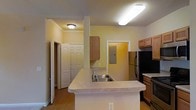 This single-floor residence is centered around an open-concept kitchen that serves as the focal point of the living space. The kitchen features a large peninsula-style island with a light-toned countertop and a stainless steel sink equipped with a chrome faucet, offering both functional workspace and a natural division between the kitchen and surrounding areas. Upper wood-toned cabinetry provides storage on both sides of the kitchen, while a black refrigerator and additional built-in appliances complete the cooking space. Overhead, a large rectangular flush-mount fluorescent fixture provides bright, even task lighting across the counters. The flooring throughout is a warm hardwood-style material that adds continuity across the space. The walls are finished in a soft yellow tone, contributing to a warm and inviting ambiance. Doorways visible from the kitchen lead to additional rooms, including what appears to be a closet with double panel doors and a secondary room, suggesting a well-connected and functional floor plan. Crown molding details along the ceiling add a subtle architectural accent to the space.