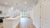 Located at 10001 Turkey Creek Rd in College Station, TX, this well-designed single-story home offers a thoughtful layout across a comfortable footprint. The open-concept kitchen and living area serves as the heart of the home, featuring light hardwood flooring, recessed lighting, and stylish pendant lights over a large center island with a white quartz countertop and undermount sink. The kitchen is equipped with stainless steel appliances, a side-by-side refrigerator, a built-in microwave, and a combination of warm wood lower cabinetry and white upper cabinets accented by a subtle tile backsplash. The living area is bathed in natural light through full-length French doors that open to outdoor views of lush greenery, complemented by a ceiling fan overhead. The bedroom is carpeted for comfort, featuring multiple windows and a ceiling fan, providing a bright and airy atmosphere. It connects directly to a dedicated closet with built-in shelving and a well-appointed bathroom. The bathroom offers a white vanity with ample cabinetry, open shelving, a tile floor, and a window for natural light. Clean white walls, modern hardware, and consistent finishes throughout give this home a fresh, move-in-ready appeal.