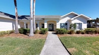 This single-story residence presents a welcoming and well-composed exterior characterized by a clean white stucco facade and a subtly decorative roofline. The front elevation is anchored by a graceful arched entryway supported by columns, leading to a bold black front door that adds a striking contrast to the light exterior. A straight concrete walkway guides visitors from the street to the entrance, flanked by neatly manicured shrubs and ground-level plantings that lend a tidy, polished curb appeal. Tall, slender palm trees rise prominently on the left side of the property, adding a tropical character consistent with a warm, coastal climate. The windows are accented with shutters, enhancing the architectural detail across the facade. Mulched garden beds frame the foundation plantings, while a well-kept lawn stretches across the front yard. The overall design conveys a sense of openness and brightness, with the wide, low-profile roofline reinforcing the home's sprawling, single-level layout. The exterior presents a cohesive, classic Florida-style aesthetic that is both inviting and understated.