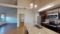 This single-floor unit showcases a thoughtfully designed open layout that blends modern finishes with functional living spaces. The kitchen serves as the centerpiece of the home, featuring rich dark wood cabinetry, speckled granite countertops, a large island with an undermount stainless steel sink, and a full suite of stainless steel appliances including a refrigerator, electric range, and built-in microwave. An elegant pendant chandelier and under-cabinet lighting illuminate the workspace, while soaring ceilings add a dramatic sense of volume throughout. The living area extends naturally from the kitchen, bathed in natural light from multiple framed windows and accented by warm hardwood-style flooring and a ceiling fan overhead. The angled roofline in the living space adds architectural character, lending the room a loft-like charm. A well-sized bedroom continues the bright, airy feel with dual windows and the same quality flooring. The bathroom is finished with classic white penny-tile flooring, a vanity with a light countertop, a shower/tub combination, and a window that frames outdoor rooftop views, filling the space with natural light. A walk-in closet with built-in shelving provides ample storage. Throughout, the clean white walls and consistent finishes create a cohesive, modern aesthetic.