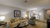 This single-floor residence offers a thoughtfully designed open-plan layout that seamlessly connects the living and dining areas, creating a cohesive and inviting atmosphere throughout. The living area is anchored by a plush cream sofa and coordinating patterned armchair, arranged around a dark oval glass-topped coffee table. Neutral beige carpeting and soft grey walls provide a warm, understated backdrop, while framed wall art adds a tasteful decorative accent. A classic table lamp and decorative accessories contribute to the room's refined character. The space flows naturally into the dining area, which is distinguished by an elegant chandelier overhead, white dining chairs, and a bright window dressed with warm golden curtains that bathe the room in natural light. A glimpse of what appears to be a kitchen or serving area is visible beyond, suggesting a well-connected floor plan designed for comfortable everyday living and entertaining. Recessed ventilation and ceiling fixtures are evenly distributed throughout, ensuring a practical and comfortable interior environment. Overall, the space presents a clean, classic aesthetic with a harmonious blend of neutral tones and carefully chosen furnishings.