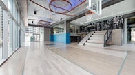 This dynamic sports and entertainment space makes an immediate impression with its expansive indoor basketball court, finished in light hardwood flooring that stretches across the main level. A regulation basketball hoop mounted on a glass backboard anchors the court, underscoring the athletic focus of the venue. Soaring ceilings overhead feature an eye-catching industrial aesthetic, with corrugated metal panels and a distinctive series of large, circular copper-toned ring sculptures suspended above — a bold artistic installation that draws the eye upward and adds a dramatic, contemporary flair.

Natural light filters in through large glass windows and doors along one side, keeping the space bright and open. A wide, modern staircase with sleek metal railings leads to an upper mezzanine level, where large-scale black-and-white photographic murals of sports legends line the walls, adding a sense of history and inspiration. A vivid blue accent wall provides a striking pop of color on the main level, complementing the overall modern industrial palette. The upper level overlooks the court below, offering an ideal vantage point for spectators. Together, the architecture, artwork, and design elements create an energetic and immersive environment built for both athletic activity and entertainment.